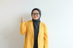 beautiful young Asian Muslim woman, wearing glasses and yellow blazer with happy expression while clenching fist photo