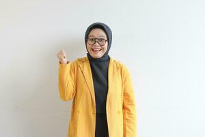 beautiful young Asian Muslim woman, wearing glasses and yellow blazer with happy expression while clenching fist photo