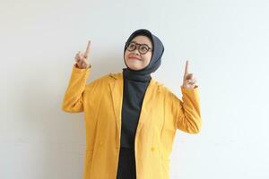 Beautiful young Asian Muslim woman in glasses, hijab and wearing yellow blazer smiling confident and happy with both hands pointing up photo