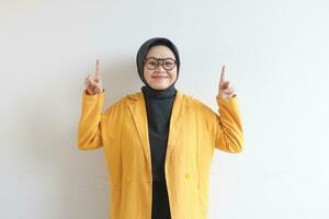 Beautiful young Asian Muslim woman in glasses, hijab and wearing yellow blazer smiling confident and happy with both hands pointing up photo