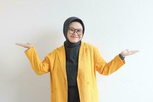 Beautiful young Asian Muslim woman in glasses, hijab and wearing yellow blazer smiling confident and happy with both hands presenting empty space photo