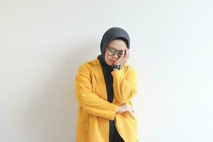 Beautiful young Asian Muslim woman, wearing glasses and yellow blazer covering 1 side of face with hand photo
