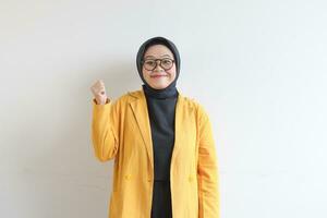 beautiful young Asian Muslim woman, wearing glasses and yellow blazer with happy expression while clenching fist photo