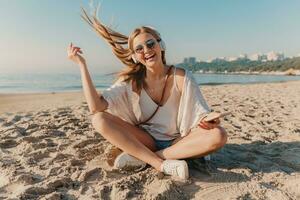 joven atractivo rubio sonriente mujer en vacaciones sentado en playa foto