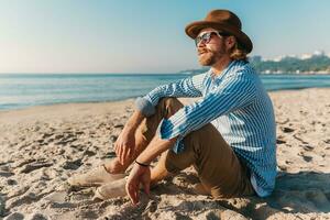 young attractive hipster man sitting on beach by sea on summer vacation photo