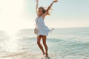 young attractive happy woman dancing turning around by sea beach in white dress vacation photo