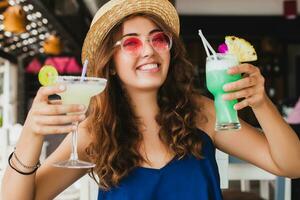 atractivo joven mujer en azul vestir y Paja sombrero vistiendo rosado Gafas de sol Bebiendo cócteles foto