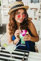 attractive young woman in blue dress and straw hat wearing pink sunglasses drinking cocktails photo