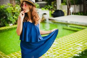 attractive young woman in blue dress and straw hat at pool of tropical spa villa photo