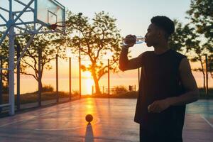 frio negro hombre haciendo Deportes en mañana, Bebiendo agua en baloncesto Corte en amanecer foto