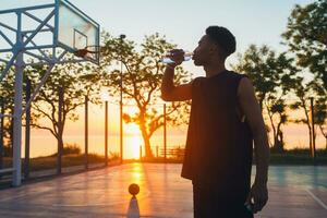 negro hombre haciendo Deportes en mañana, Bebiendo agua en baloncesto Corte en amanecer foto