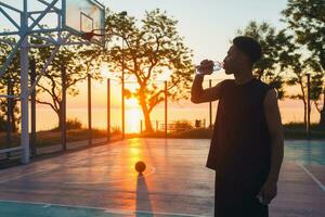 black man doing sports in morning, drinking water on basketball court on sunrise photo