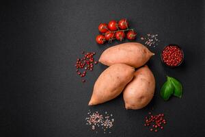 Fresco grande rosado dulce patata tubérculos con Tomates y especias en un oscuro antecedentes foto