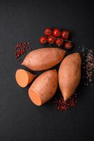 Fresco grande rosado dulce patata tubérculos con Tomates y especias en un oscuro antecedentes foto