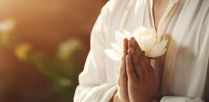 A Buddhist Man in a White Robe is Praying and Holding a Blooming Lotus Flower in his Hands on his Chest. Warm Natural Light Shading on. photo