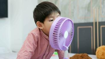 asiatico ragazzo dire bugie su il materasso su un' caldo giorno giocando con un' portatile fan felicemente video