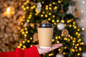 A disposable cup with a lid stands on a woman's hand near a Christmas tree with a garland photo