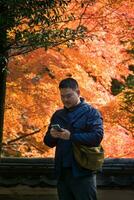 asiático hombre mientras utilizando móvil teléfono debajo arce árbol hojas durante otoño con color cambio en hoja en naranja amarillo y rojo foto