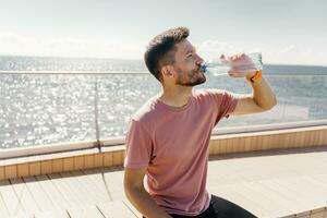 Drinks water from a bottle, rest break. Athlete runner training in sportswear. A sporty man running a lot. Man running in the city fresh air. photo