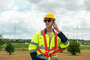 un inteligente ingeniero con protector casco en cabeza, utilizando teléfono inteligente a eléctrico turbinas campo foto