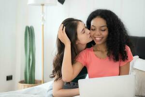 Happy lesbian couple using laptop on bed at home. Young multiethnic lesbian couple using laptop together in bedroom. Technology and love concept photo