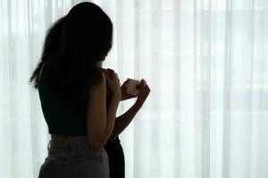 Both of cheerful young women drinking coffee at home holding coffee cups and chatting behind the white curtains of the living room where light shines photo