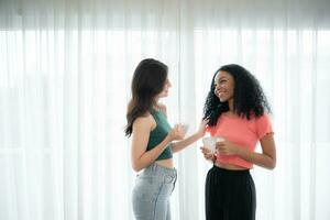 Both of cheerful young women drinking coffee at home holding coffee cups and chatting behind the white curtains of the living room where light shines photo