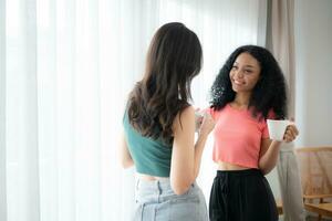 Both of cheerful young women drinking coffee at home holding coffee cups and chatting behind the white curtains of the living room where light shines photo