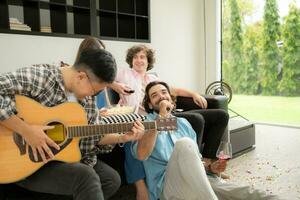 multiétnico grupo de amigos teniendo divertido jugando guitarra y canto juntos a hogar foto