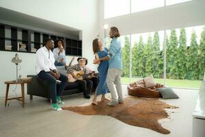 Group of multiethnic friends having fun at party by playing guitar, singing and dance together at home. photo