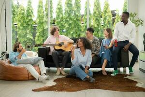 grupo de multiétnico amigos teniendo divertido a fiesta por jugando guitarra y canto juntos a hogar. foto