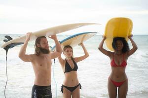 posterior ver de dos mujer y joven hombre participación tablas de surf en su cabezas y caminar dentro el mar a navegar foto