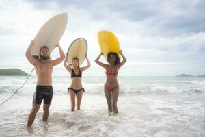 Rear view of two women and young man holding surfboards on their heads and walk into the sea to surf photo