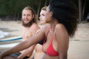 Group of multiracial friends sitting on surfboards on sandy beach photo