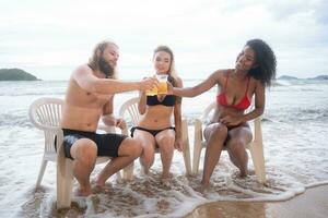 Multi-ethnic group of friends sitting on chairs with drinks on the beach photo