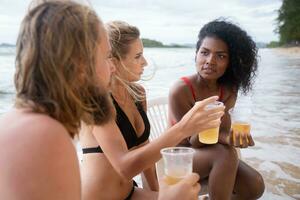 Multi-ethnic group of friends sitting on chairs with drinks on the beach photo