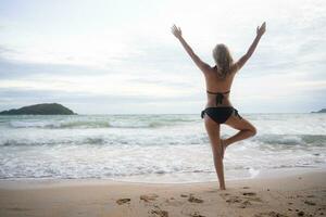 Young woman in black bikini practicing yoga on the beach at sunset. photo