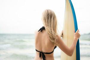 retrato de sonriente joven mujer en pie con tabla de surf a el playa foto