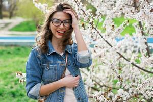 Sexy woman in jeans jacket, red full lips, wavy hairstyle posing near flower tree in spring day. photo