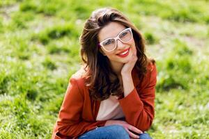 Outdoor portrait of attractive woman with eyeglasses, red jacket, wavy hairstyle sitting on grass. photo