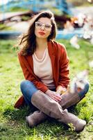 elegante mujer en casual primavera atuendo sentado en césped en soleado calentar parque , disfrutando descanso día. foto