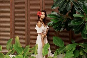 Graceful Asian woman with perfect skin  and hibiscus flower in hairs posing over wood wall and tropical plants. Summer fashion portrait. photo