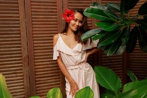 Graceful Asian woman with perfect skin  and hibiscus flower in hairs posing over wood wall and tropical plants. Summer fashion portrait. photo