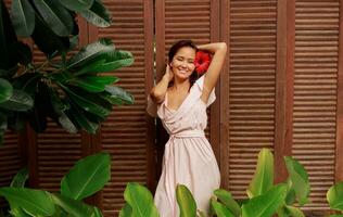 agraciado asiático mujer con Perfecto piel y hibisco flor en pelos posando terminado madera pared y tropical plantas. verano Moda retrato. foto
