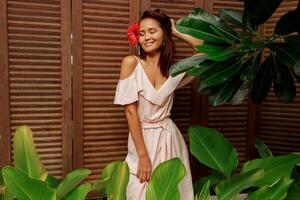 Graceful Asian woman with perfect skin  and hibiscus flower in hairs posing over wood wall and tropical plants. Summer fashion portrait. photo