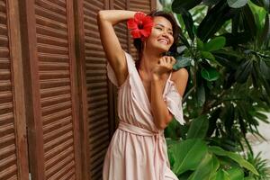 Graceful Asian woman with perfect skin  and hibiscus flower in hairs posing over wood wall and tropical plants. Summer fashion portrait. photo