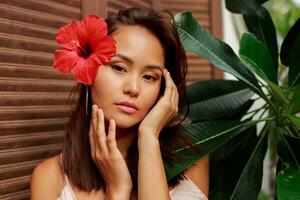 agraciado asiático mujer con Perfecto piel y hibisco flor en pelos posando terminado madera pared y tropical plantas. verano Moda retrato. foto