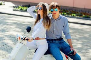 Fashionable  couple posing  on the street, siting on scooter , wearing stylish casual clothes, bright sunglasses. Smiling  handsome guy and his pretty girlfriend hugging.  Man holding retro camera. photo