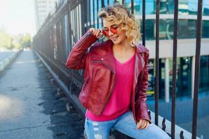 Happy  blond  woman posing on modern  streets, drinking coffee  or cappuccino.  Stylish autumn outfit,  leather jacket and knitted sweater. Urban background. Pink sunglasses. photo