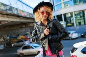 Happy  blond  woman posing on modern  streets, drinking coffee  or cappuccino.  Stylish autumn outfit,  leather jacket and knitted sweater. Urban background. Pink sunglasses. photo